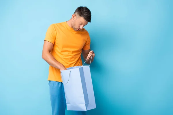 Überraschter Mann im T-Shirt blickt auf Einkaufstasche auf blauem Grund — Stockfoto