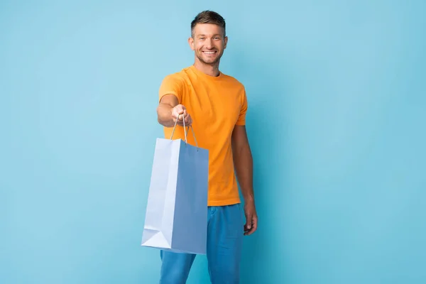 Fröhlicher Mann im T-Shirt mit ausgestreckter Hand hält Einkaufstasche auf blau — Stockfoto