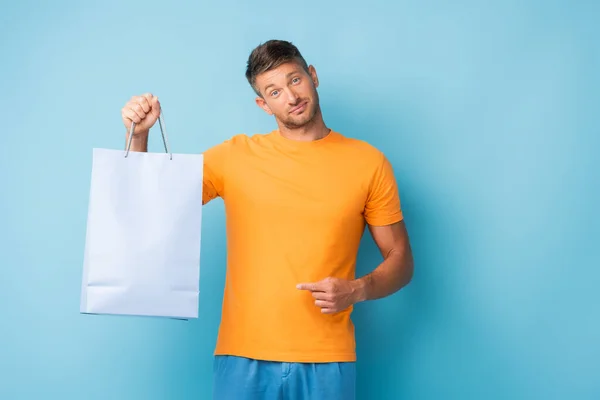 Uomo confuso in t-shirt punta con dito shopping bag su blu — Foto stock