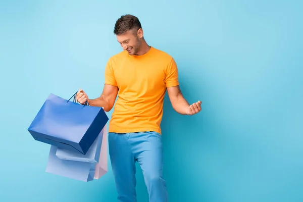 Felice uomo in t-shirt che tiene borse della spesa su blu — Foto stock