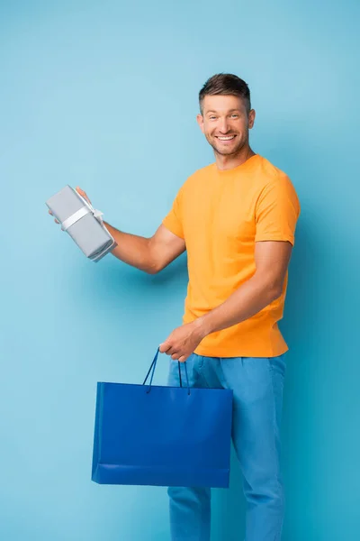 Cheerful man in t-shirt holding shopping bag and present on blue — Stock Photo