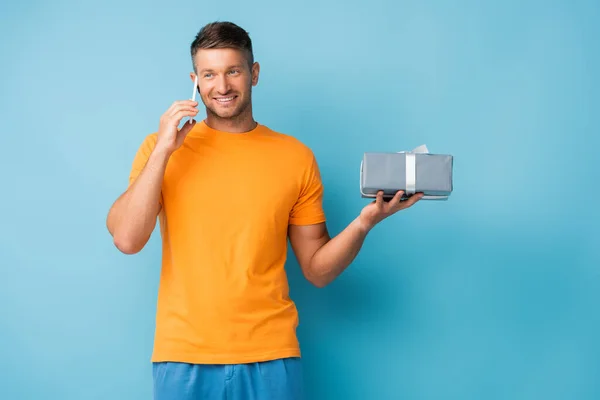 Felice uomo in t-shirt contenente scatola regalo e parlando su smartphone su blu — Foto stock