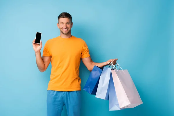 Glücklicher Mann mit Einkaufstaschen und Smartphone mit leerem Bildschirm auf blauem Hintergrund — Stockfoto
