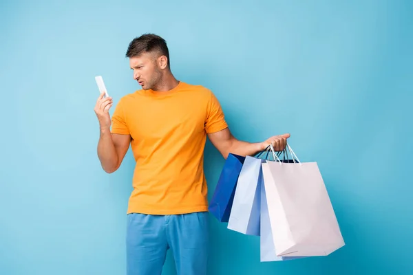 Homem feliz segurando sacos de compras e olhando para o smartphone em azul — Fotografia de Stock