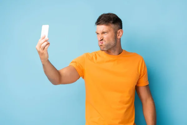 Hombre insatisfecho en camiseta sosteniendo el teléfono inteligente y tomando selfie en azul - foto de stock