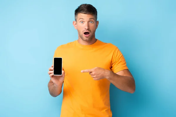 Hombre sorprendido en camiseta apuntando con el dedo en el teléfono inteligente con pantalla en blanco en azul - foto de stock