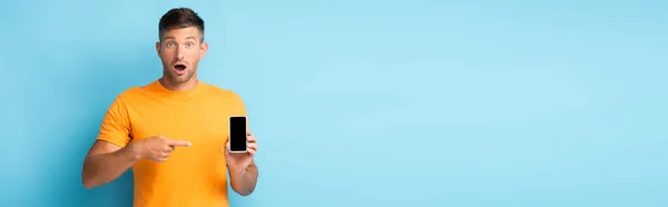 Шокований чоловік у футболці, що вказує пальцем на смартфон з порожнім екраном на синьому, банер — стокове фото