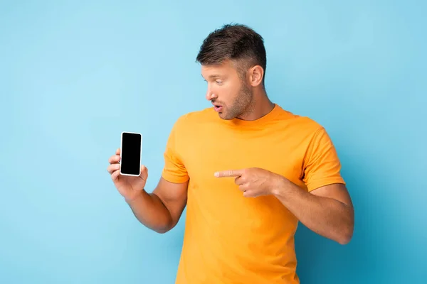 Uomo sorpreso in t-shirt che punta con il dito verso smartphone con schermo bianco su blu — Foto stock