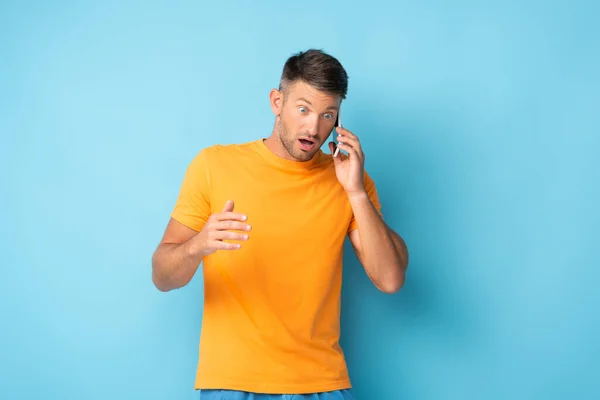 Homme bouleversé homme en t-shirt parler sur smartphone sur bleu — Photo de stock