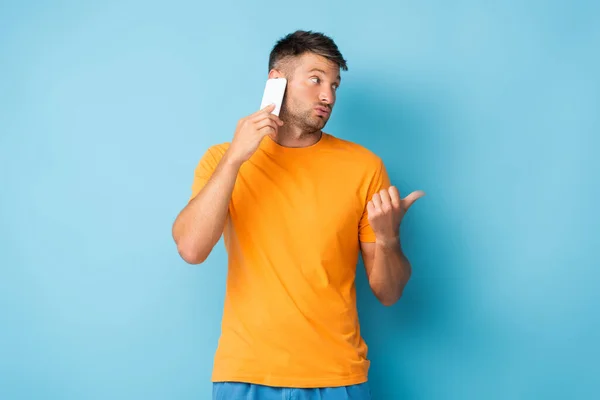 Homem em t-shirt falando no smartphone e apontando com o polegar no azul — Fotografia de Stock