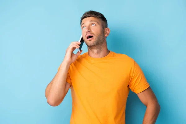 Hombre en camiseta hablando en el teléfono inteligente y mirando hacia arriba en azul - foto de stock