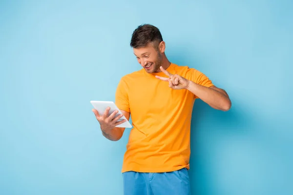 Glücklicher Mann im T-Shirt mit Friedenszeichen, während er digitales Tablet auf blau benutzt — Stockfoto