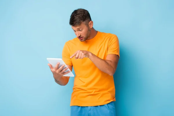 Homem descontente em t-shirt apontando com o dedo para tablet digital em azul — Fotografia de Stock