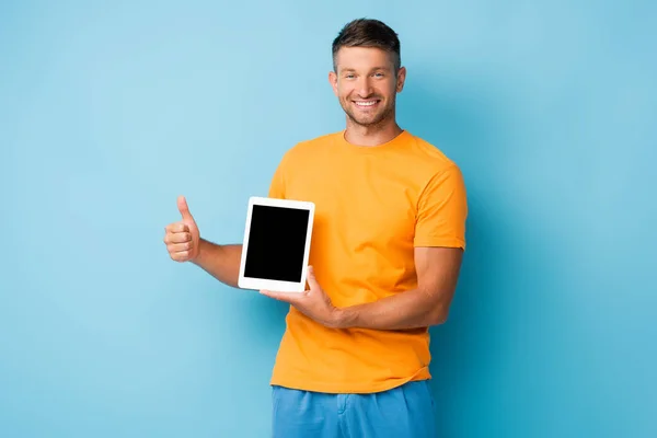Glücklicher Mann im T-Shirt hält digitales Tablet mit leerem Bildschirm und zeigt Daumen nach oben auf blau — Stockfoto