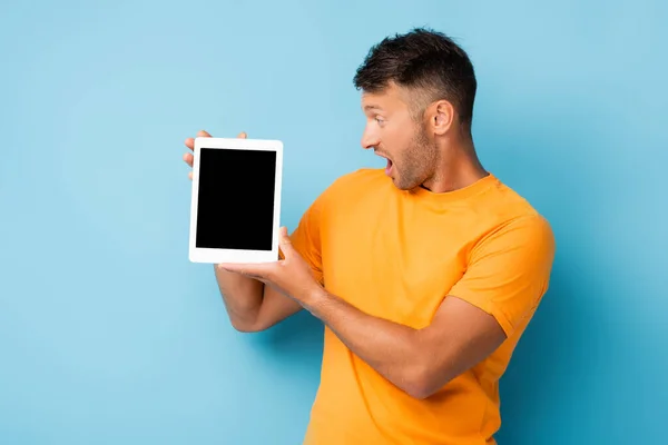 Überraschter Mann im T-Shirt blickt auf digitales Tablet mit leerem Bildschirm auf blauem Hintergrund — Stockfoto