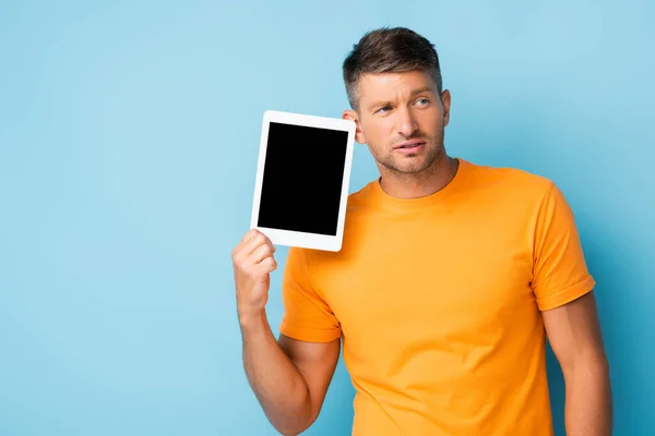 Спантеличений чоловік у футболці тримає цифровий планшет з порожнім екраном на синьому — стокове фото