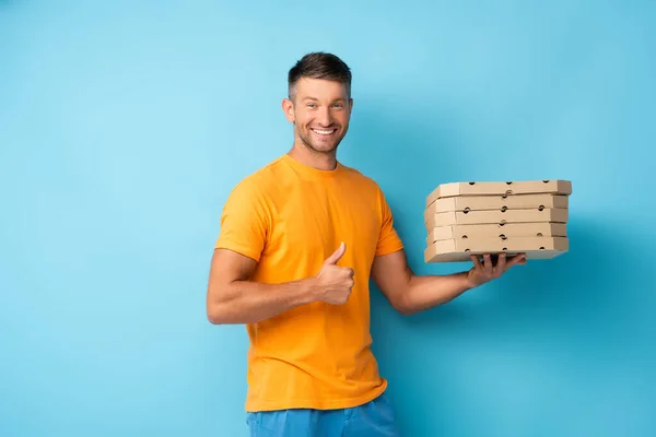 Hombre feliz en camiseta sosteniendo cajas de pizza de cartón y mostrando el pulgar hacia arriba en azul - foto de stock