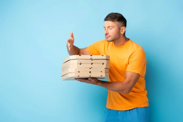 Hombre con los ojos cerrados oliendo pizza mientras sostiene cajas en azul - foto de stock