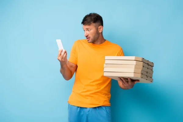 Homem em t-shirt segurando caixas de pizza de papelão e olhando para smartphone em azul — Fotografia de Stock