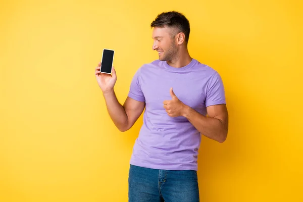Hombre feliz sosteniendo teléfono inteligente con pantalla en blanco y mostrando el pulgar hacia arriba en amarillo - foto de stock