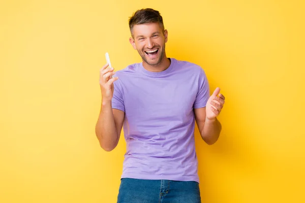 Excited man holding smartphone and laughing on yellow — Stock Photo