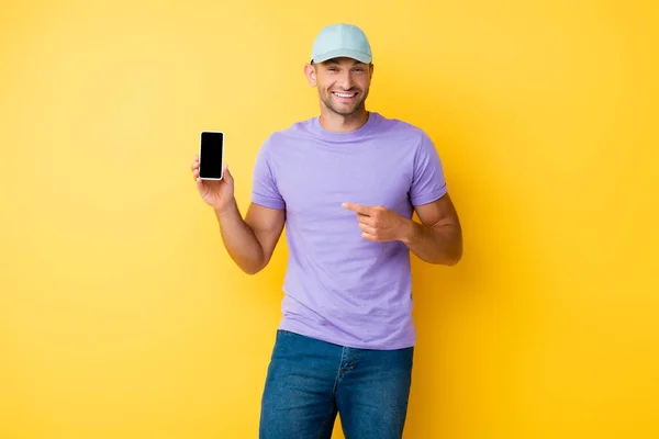 Щасливий чоловік у блакитній шапці, вказуючи пальцем на смартфон з порожнім екраном на жовтому — стокове фото