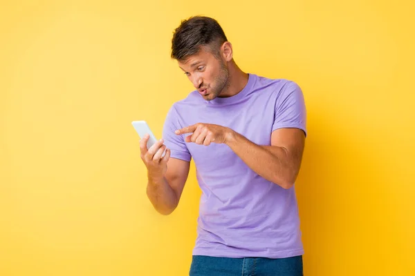 Hombre disgustado haciendo pucheros labios mientras que señala con el dedo en el teléfono inteligente en amarillo - foto de stock