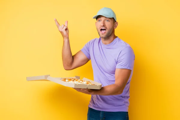 Mann mit blauer Mütze gestikuliert, während er Schachtel mit leckerer Pizza auf gelb hält — Stockfoto