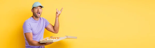 Человек в синей кепке жестикулируя, держа коробку с вкусной пиццей на желтом, баннер — стоковое фото