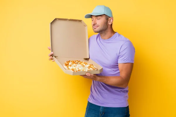 Homem satisfeito em azul cap cheirando pizza saborosa em caixa de papelão em amarelo — Fotografia de Stock