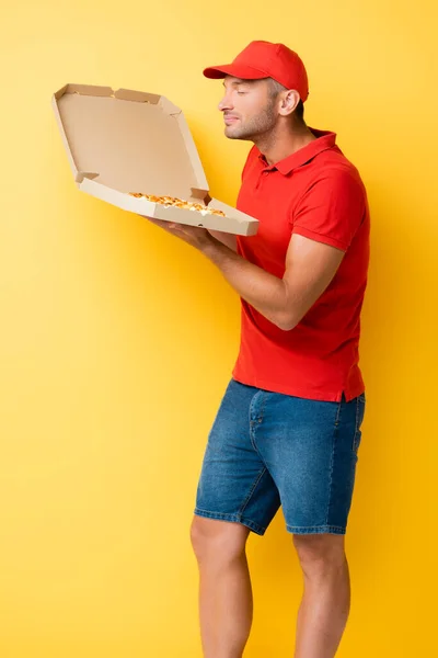 Uomo di consegna contento in tappo rosso profumando gustosa pizza in scatola di cartone su giallo — Foto stock