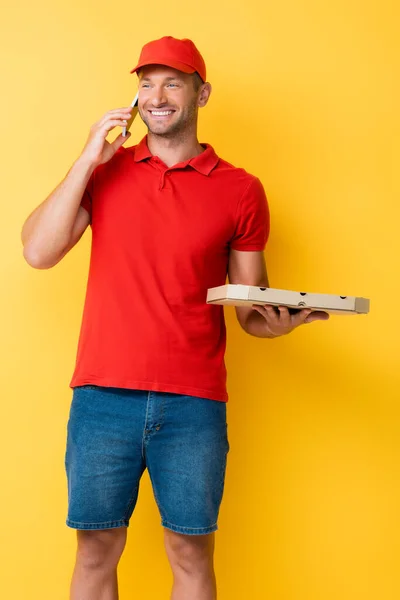 Heureux livreur dans la boîte de maintien chapeau rouge avec pizza savoureuse et parler sur smartphone sur jaune — Photo de stock