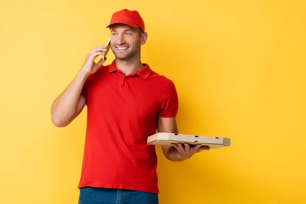 Smiling delivery man in red cap holding box with tasty pizza and talking on smartphone on yellow — Stock Photo