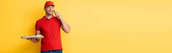 Repartidor sonriente en caja de sujeción de tapa roja con sabrosa pizza y hablando en el teléfono inteligente en amarillo, bandera - foto de stock