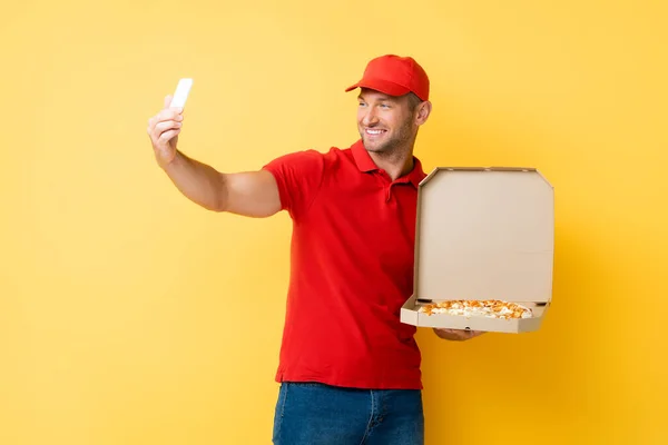 Repartidor sonriente en caja de sujeción de tapa roja con sabrosa pizza y tomando selfie en amarillo - foto de stock