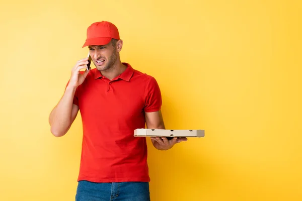 Displeased delivery man in red cap holding box with tasty pizza and talking on smartphone on yellow — Stock Photo