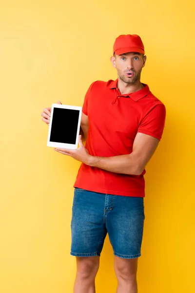 Schockierter Zusteller mit roter Mütze hält digitales Tablet mit leerem Bildschirm auf gelb — Stockfoto