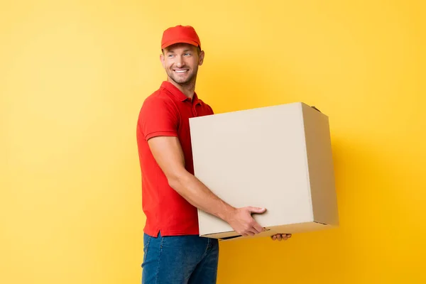 Lächelnder Lieferant in roter Mütze mit Karton auf gelb — Stockfoto