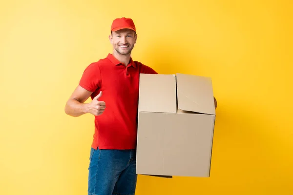 Homem de entrega feliz em tampa vermelha segurando caixa de papelão e mostrando polegar para cima no amarelo — Fotografia de Stock