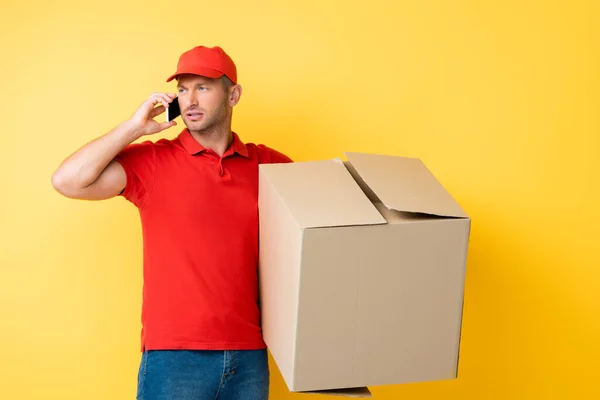 Repartidor en tapa roja sosteniendo caja de cartón y hablando en el teléfono inteligente en amarillo — Stock Photo