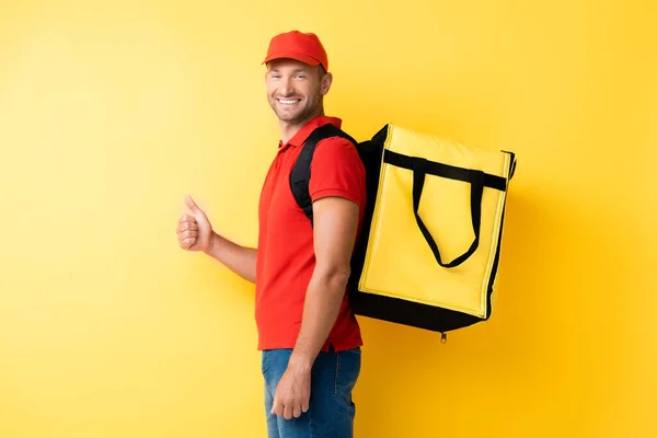 Cheerful delivery man carrying backpack with order and showing thumb up on yellow — Stock Photo
