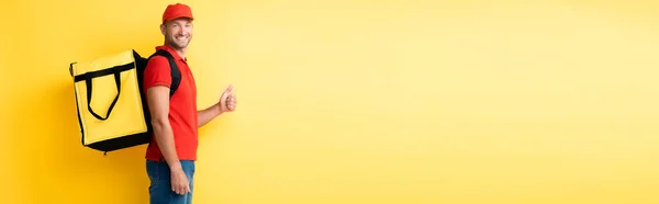 Cheerful delivery man carrying backpack with order and showing thumb up on yellow, banner — Stock Photo