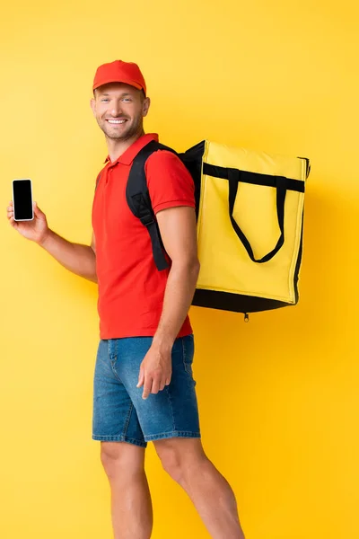Cheerful delivery man carrying backpack and holding smartphone with blank screen on yellow — Stock Photo