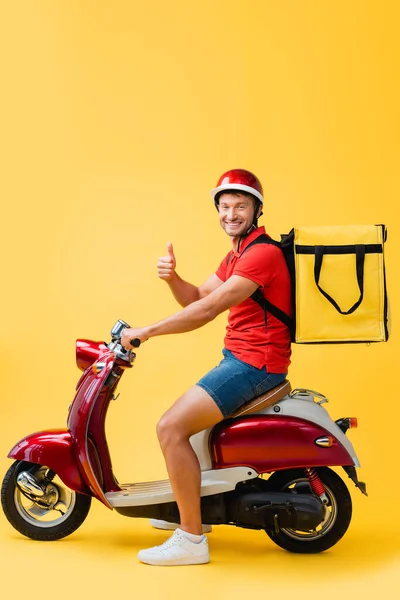 Happy delivery man in helmet and backpack riding red scooter while showing thumb up on yellow — Stock Photo