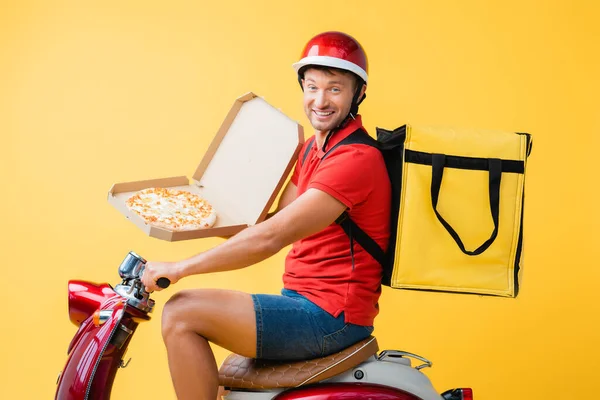 Homem de entrega feliz com mochila montando scooter vermelho e segurando pizza na caixa de papelão no amarelo — Fotografia de Stock