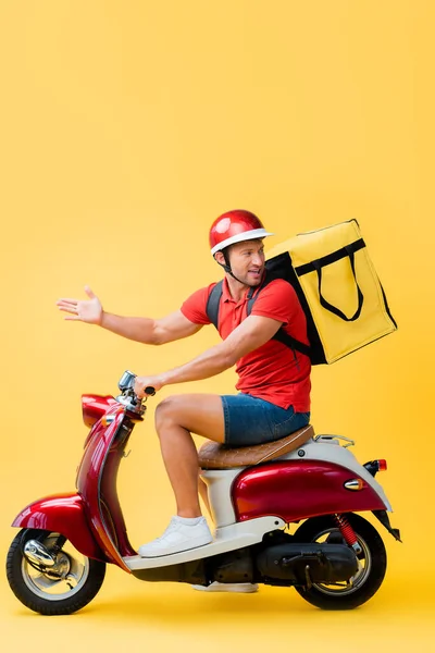 Homem de entrega descontente com a mochila gesticulando ao andar de scooter no amarelo — Fotografia de Stock