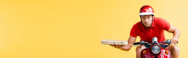 Concentrated delivery man in helmet riding red scooter and holding pizza box isolated on yellow, banner — Stock Photo