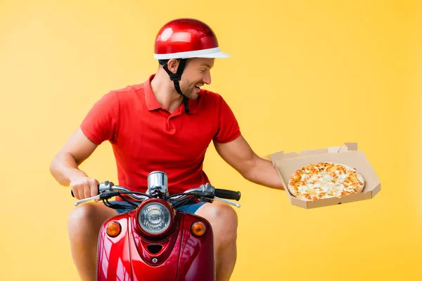 Felice uomo di consegna in casco cavalcando scooter rosso e tenendo la pizza in scatola di cartone isolato su giallo — Foto stock