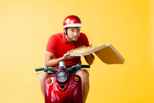 Delivery man in helmet riding red scooter and smelling pizza in carton box on yellow — Stock Photo