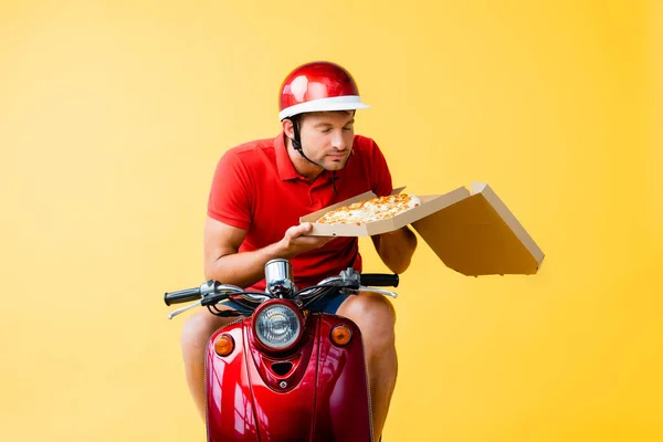 Homem de entrega no capacete montando scooter vermelho e cheirando pizza na caixa no amarelo — Fotografia de Stock